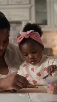 Vertical of young Black mom teaching her toddler daughter how to draw on paper with crayons and kissing her