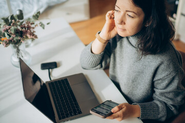 Young woman with dark hair, wearing a cozy gray sweater, is engaged in a phone conversation while working on a laptop at a bright, modern workspace with natural light