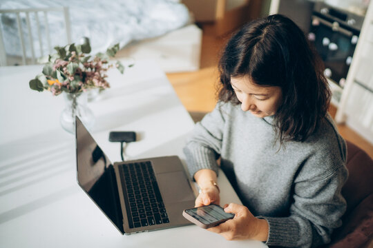 Young woman with dark hair, wearing a cozy sweater, is using a smartphone while sitting at a desk with a laptop, surrounded by a bright and inviting home office atmosphere