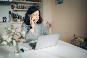 Woman talking on the phone while working on a laptop at a cozy kitchen table during the day