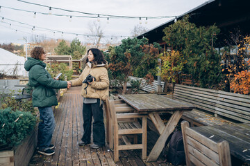 Two women, one with curly hair and green jacket, shake hands in a cozy outdoor garden setting, surrounded by wooden furniture and vibrant plants, capturing a moment of connection