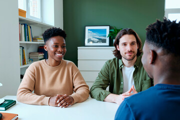 Obraz premium Young Black woman and young man sitting at desk listening to young Black man during study session in room with bookshelves and framed picture