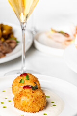 Close-up of two crispy golden arancini served on a white plate with a creamy sauce and microgreens garnish. A wine glass and other appetizers are blurred in the background.