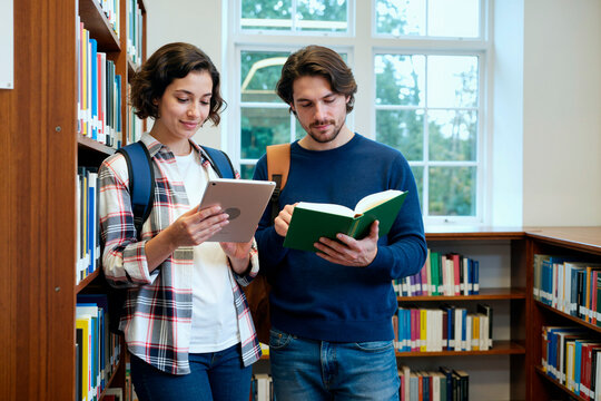 Young woman holding digital tablet standing next to young man reading book in library, both wearing backpacks and studying together near bookshelves