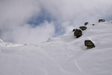 Snow-covered alpine slope with scattered rocks, strong wind patterns, and fog moving around a rugged mountain. Atmospheric winter landscape in cold high-altitude conditions.