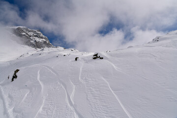 Snow-covered alpine slope with scattered rocks, strong wind patterns, and fog moving around a rugged mountain. Atmospheric winter landscape in cold high-altitude conditions.