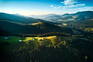 Fototapeta premium Aerial view of picturesque valley with rolling hills, lush forests, and scattered houses. Sunlight bathes landscape in warm glow, highlighting vibrant greens and creating serene atmosphere.