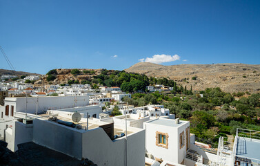 Panoramic view of traditional white Mediterranean houses and green valley beneath rocky hills under clear blue summer sky in a Greek island village.