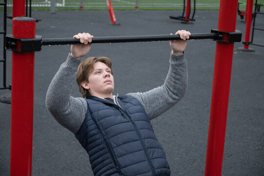 Young man performing pull-up exercise at an outdoor gym, holding onto a bar. Strength training in public fitness area. Outdoors - Powered by Adobe