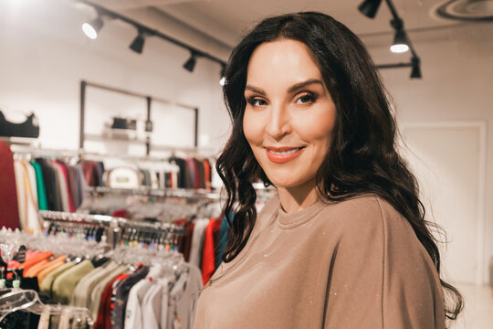 Confident woman with long dark hair smiles while standing in a clothing store, surrounded by colorful garments on racks, showcasing a vibrant shopping atmosphere - Powered by Adobe