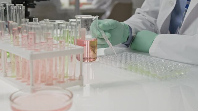 Close-up shot of unrecognizable scientist in lab coat and gloves pipetting liquid into microplate in clean laboratory desk setup