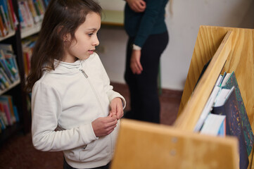 Young Girl Exploring Books in School Library While a Teacher Oversees Learning