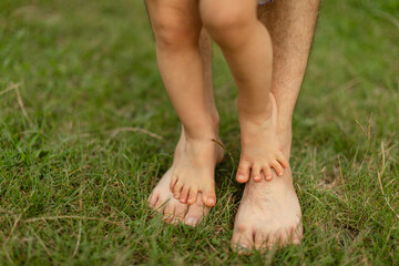 Close-up of small bare feet standing on top of adult feet on green grass outdoors.