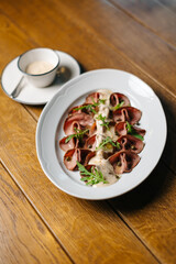 Appetizing beef slices with creamy sauce and arugula served on a white plate on wooden table, with a cup of sauce in the background. Elegant restaurant food presentation.