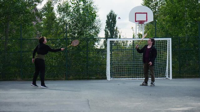 Two People playing shuttlecock kicking game on a grey concrete court in an outdoor park area during daylight