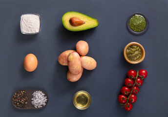 Various fresh ingredients are neatly arranged on a dark surface, including an avocado, potatoes, cherry tomatoes, egg, and seasonings. The vibrant colors invite creativity in cooking.