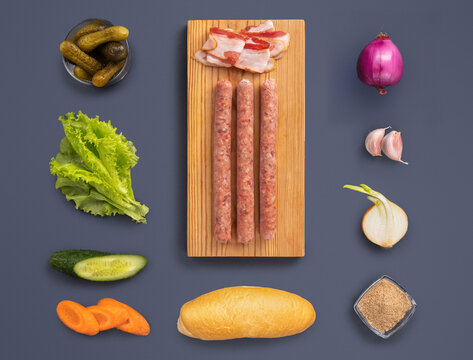 On a wooden board, sausages lay between fresh vegetables like lettuce, cucumber, and pickles. Slices of bacon, onion, garlic, and a bread roll add delicious flavor options for a sandwich.