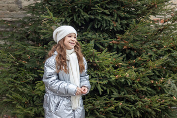 Smiling girl in silver puffer jacket and white scarf beside evergreen trees outdoors © Nataliia