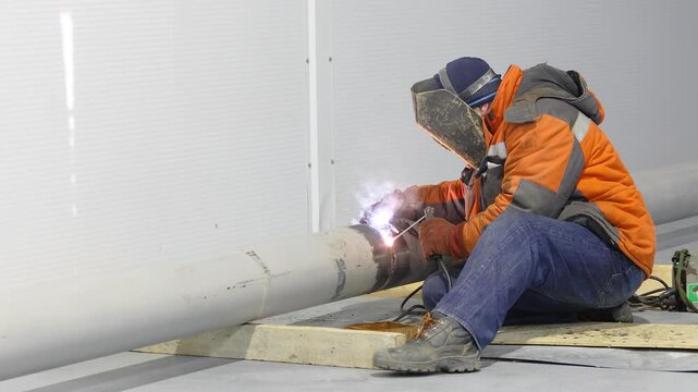 Welder welding a metal pipe or pipeline. Close-up of welding work indoor. Welder at a construction site. Welding work inside a large industrial or commercial building. Welding seam, sparks, flash.