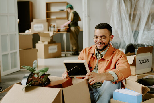 Young adult man sitting among moving boxes smiling while unpacking framed photo, young adult woman in background organizing items during home renovation and relocation