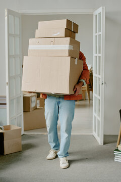 Caucasian young adult man carrying large stack of cardboard boxes while moving into new home, standing in doorway with additional moving boxes visible in background