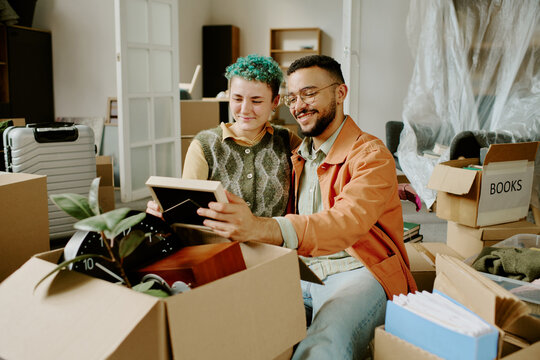 Woman and young adult man unpacking moving boxes together, smiling and looking at framed photo, surrounded by packed belongings during home renovation - Powered by Adobe
