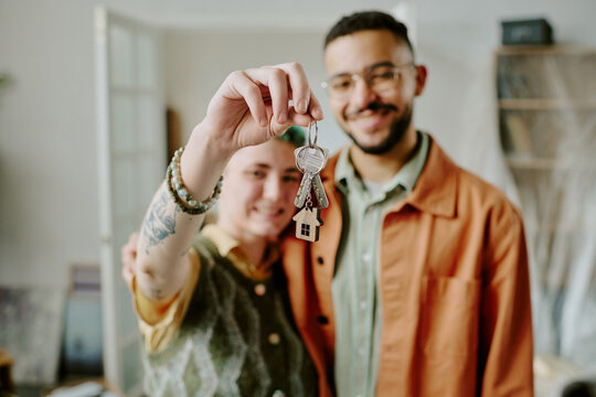 Young adult woman and man smiling and holding house keys together in home interior, showing keys toward camera, couple moving and doing renovations