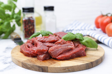 Pieces of raw beef meat, basil and ingredients on white marble table, closeup