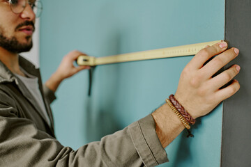Young adult man measuring wall with tape measure during home renovation, focusing on precise alignment, partial face and hands visible, engaged in moving and improvement