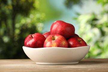 Ripe red apples in bowl on wooden table against blurred green background, closeup