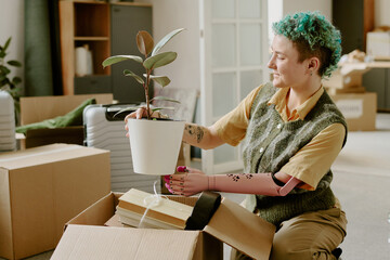 Young adult Caucasian woman with prosthetic arm unpacking cardboard box and placing potted plant inside modern home during moving and renovation process, surrounded by packed belongings