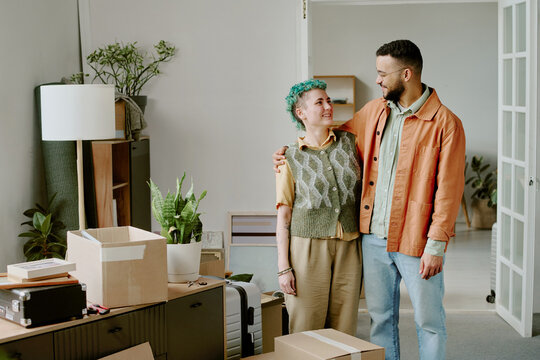 Caucasian woman with short green hair and young adult man standing together smiling, embracing each other while unpacking moving boxes in new home during renovation