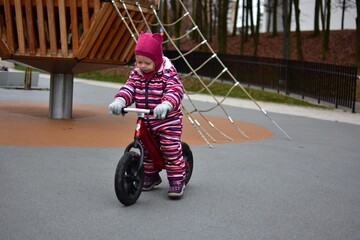 Toddler girl in a striped winter suit riding a small balance bike on a modern playground with climbing nets and wooden structures. Active outdoor play on a cool autumn day.
