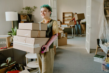 Young adult Caucasian woman with short curly hair carrying stack of cardboard boxes while smiling, young adult man in background moving boxes during home renovation 