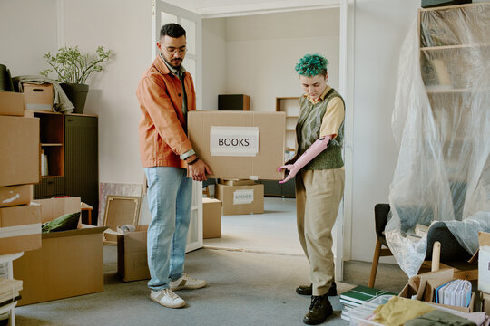 Young adult man and young adult woman with prosthetic arm carrying cardboard box labeled books while moving and renovating apartment surrounded by packed belongings