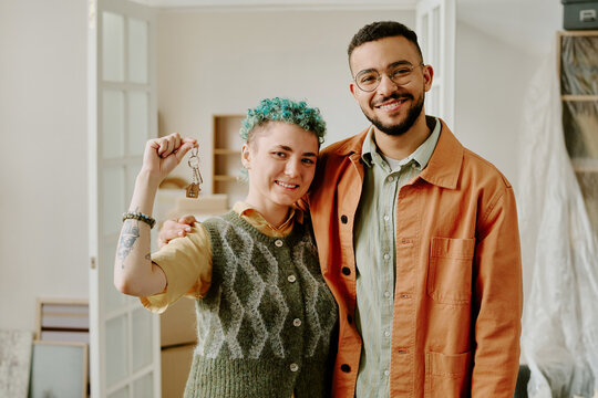 Young adult Caucasian woman with short curly blue hair holding keys standing next to young adult man smiling, couple posing together during moving or home renovation