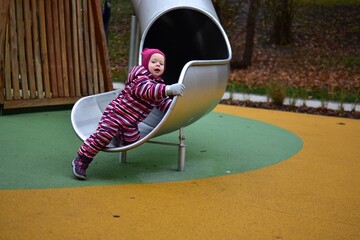 Little girl in a striped winter suit holding onto a metal slide and looking back with curiosity. Colorful soft flooring and wooden playground elements in the background.
