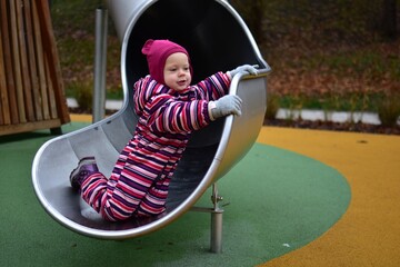 Little girl in a striped winter suit holding onto a metal slide and looking back with curiosity. Colorful soft flooring and wooden playground elements in the background.