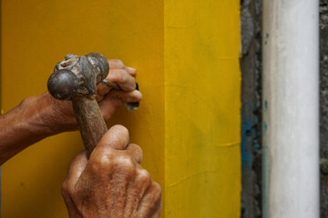 a pair of wrinkled hands are chiseling a hole in a yellow wall outdoor manual work process with...