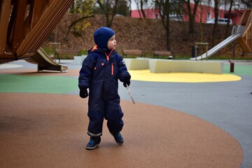 Little boy in a blue winter overall walking around a colorful outdoor playground, holding a stick and exploring the play area during a chilly autumn day.