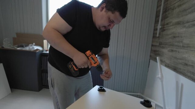 Professional furniture assembler working indoors, using a cordless electric drill to screw parts together while putting up a new kitchen cabinet during a home improvement and renovation project.