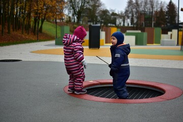 Two toddlers dressed in warm winter suits standing near a sunken playground trampoline on a colorful outdoor play area, enjoying autumn weather together. Siblings having fun outside.
