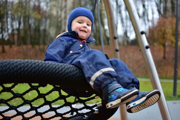 Smiling toddler boy in a blue winter suit sitting on a large rope swing at a modern playground. Autumn trees and soft play surfaces form a cozy outdoor scene.