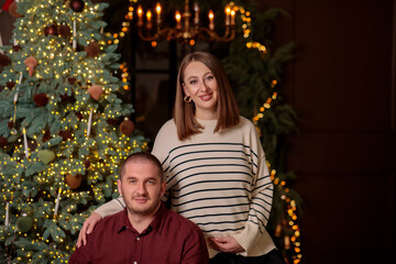 Happy pregnant couple smiling and embracing near a brightly lit christmas tree, celebrating the holiday season and expecting a baby