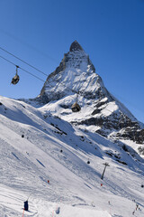 Ski lift gondolas passing above the snowy Matterhorn peak on a clear winter day. iconic swiss alpine scenery with dramatic mountain view and bright blue sky.