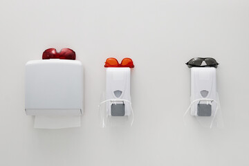 Three wall mounted dispensers with colored safety glasses arranged above, representing dental...