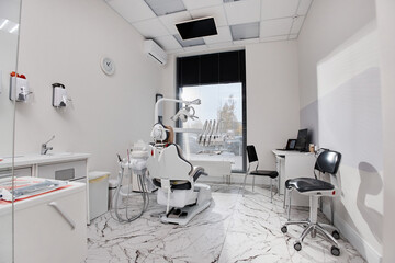 Empty dental office showing modern dental chair and equipment arranged in clean clinical setting, large window providing natural light, workspace prepared for patient appointment