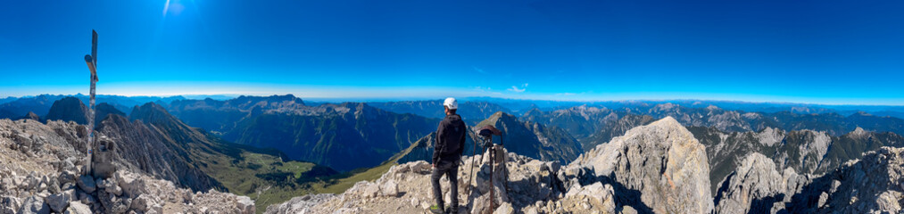 A climber stands between the summit cross and bell on Jof di Montasio, admiring the panoramic view...