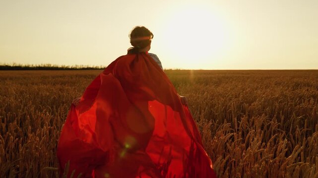 Happy child girl play superheroes in nature. Kid in cape play heroes. Child girl play superhero in red cape on wheat field, children's dream. Child hero in red cape into sun. Childhood dream red cloak