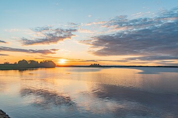 Serene sunset over a calm, reflective lake Orange and pink sky gradient with scattered clouds Mirrored colors on water surface Subtle ripples Distant land with vegetation No man-made structures